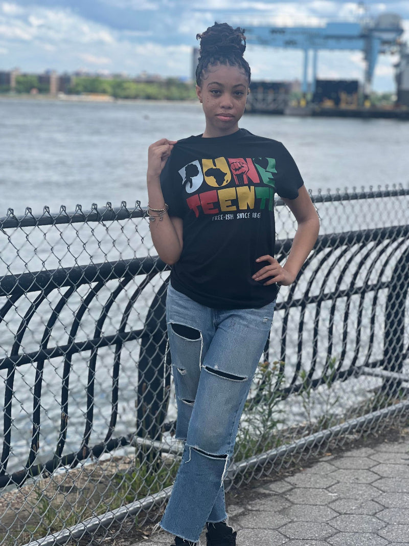A person standing by a fence wearing a graphic t-shirt with Juneteenth-themed design featuring an African American woman with a powerful afro and graffiti-style artwork.