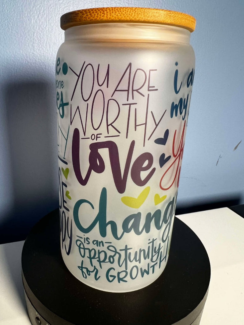 A frosted glass motivational cup with colorful text and designs, displaying affirmations such as 'You are worthy of love' and 'You are loved', with a bamboo lid on top and a straw included.