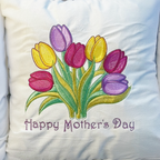 A square-shaped embroidered pillow with a design of pink, yellow, and lavender tulips and the text 'Happy Mother's Day'. The pillow is displayed in a field of real tulips.
