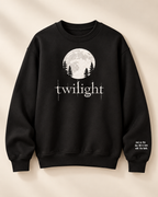 A black crewneck sweatshirt featuring a white graphic of a full moon, pine trees, and the word 'twilight' on the chest, with small white text on the right sleeve, displayed against a warm beige background.