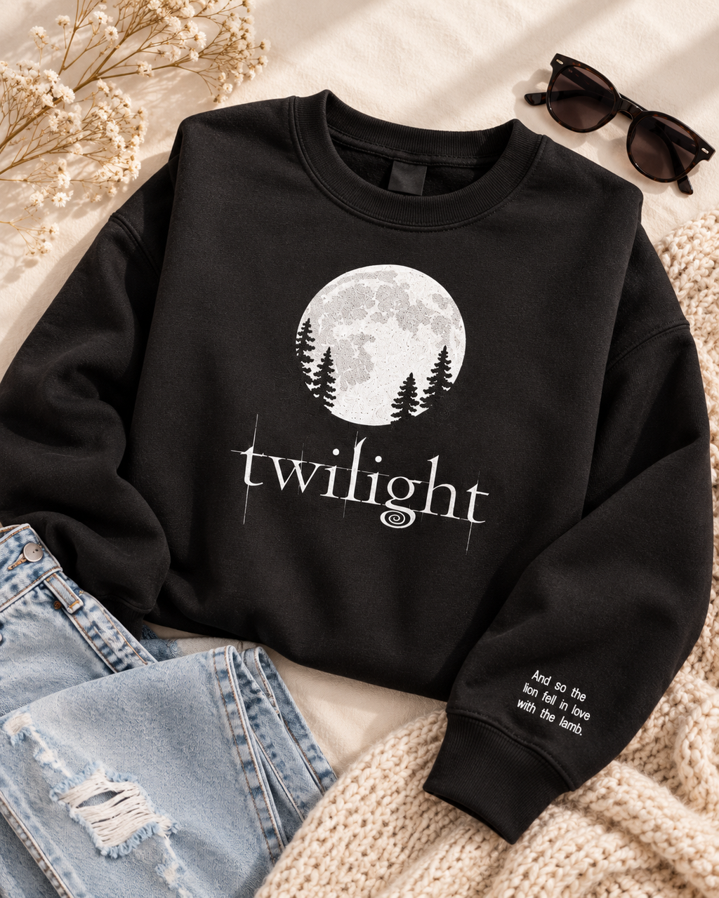 A black crewneck sweatshirt with a white graphic of a full moon and pine trees, with the word 'twilight' printed below. The sweatshirt is styled in a flat lay with light blue jeans, a beige knit blanket, sunglasses, and dried flowers on a light beige background.