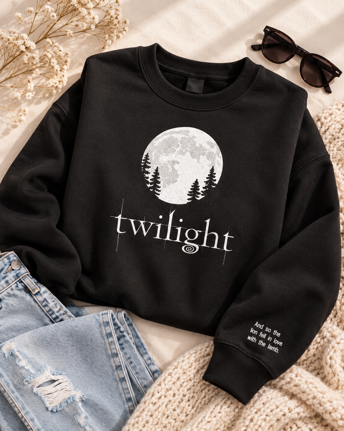 A black crewneck sweatshirt with a white graphic of a full moon and pine trees, with the word 'twilight' printed below. The sweatshirt is styled in a flat lay with light blue jeans, a beige knit blanket, sunglasses, and dried flowers on a light beige background.