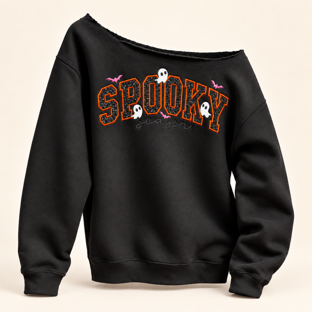 A black off-the-shoulder sweatshirt featuring a 'Spooky Season' graphic on the chest with orange glitter-style lettering, white ghosts, and pink bats, set against a light beige background.