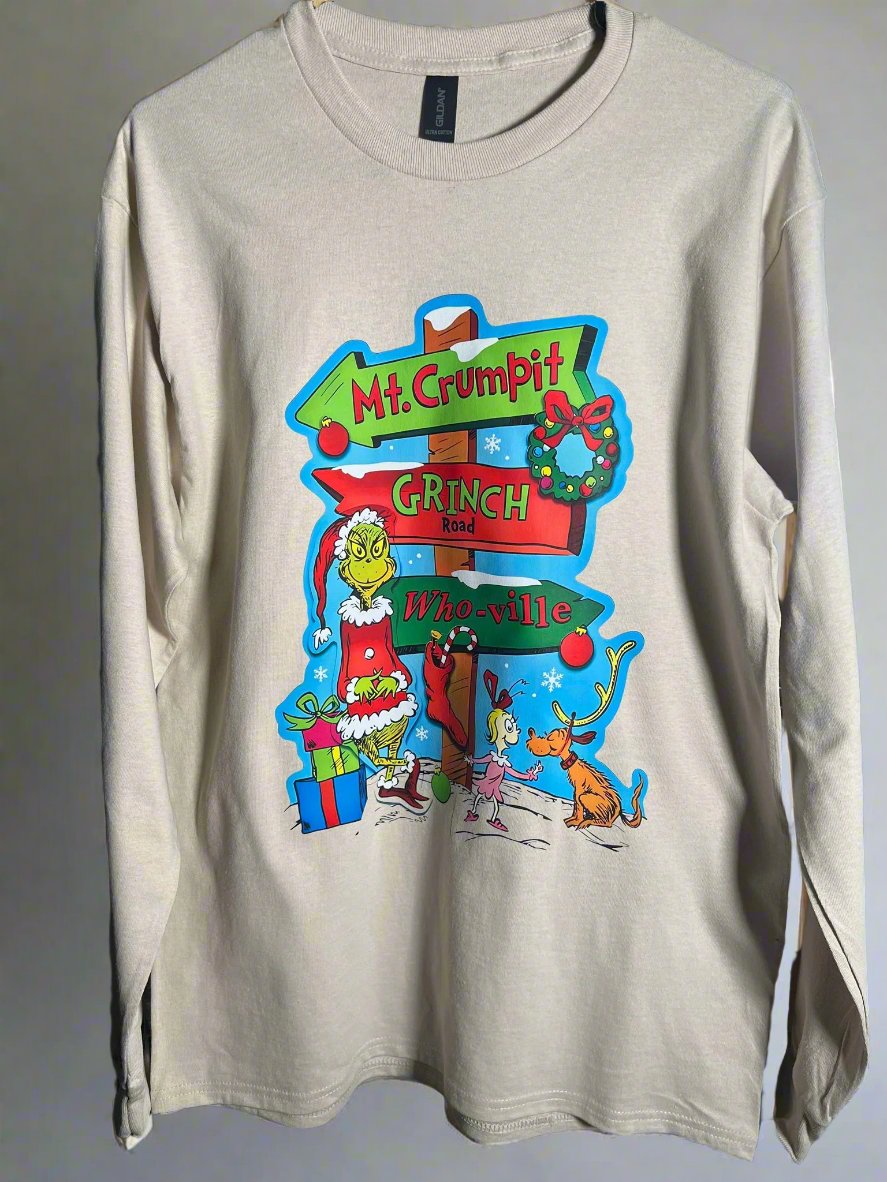A cream-colored t-shirt with a graphic design featuring the character 'The Grinch' and a whimsical road sign pointing to Mt. Crumpit and Whoville.