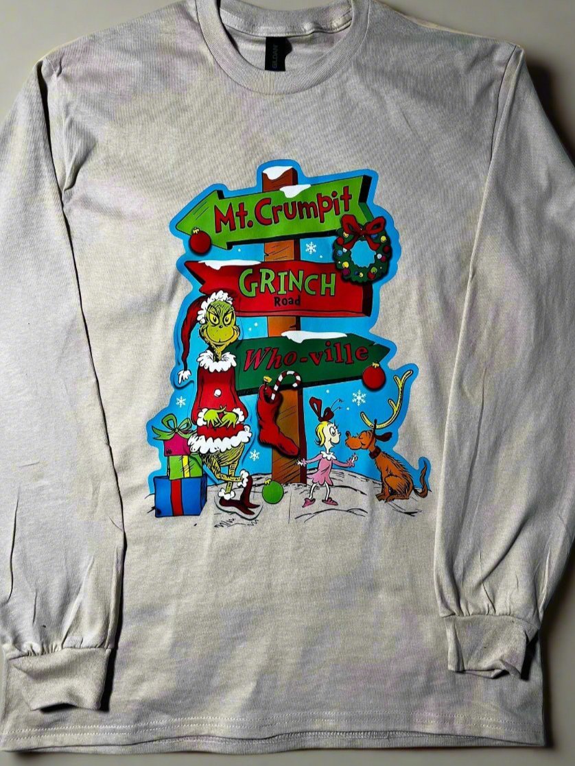 A cream-colored t-shirt with a graphic design featuring the character 'The Grinch' and a whimsical road sign pointing to Mt. Crumpit and Whoville.