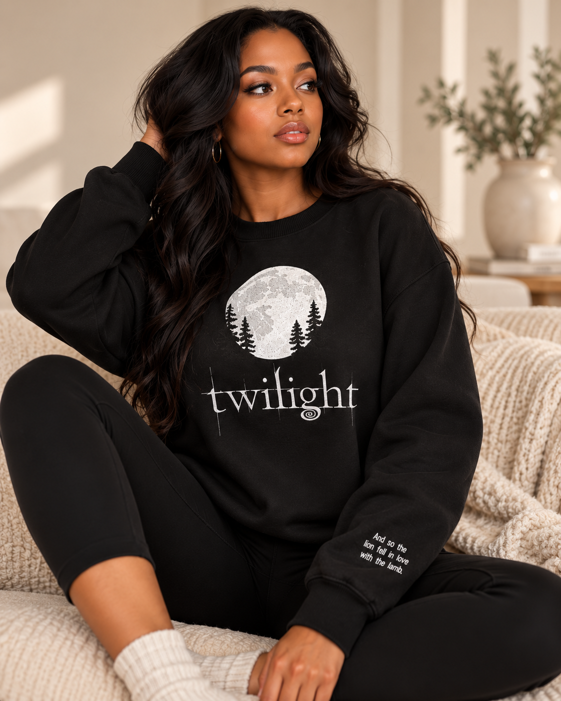 A woman sits on a cream knit sofa wearing a black crewneck sweatshirt with a white moon and pine tree graphic and the word 'twilight' printed across the front, paired with black leggings.