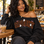 A woman sitting at a cafe table wearing a black off-the-shoulder sweatshirt with 'SPOOKY season' written in orange and white glitter-style lettering, with small ghost and bat graphics.