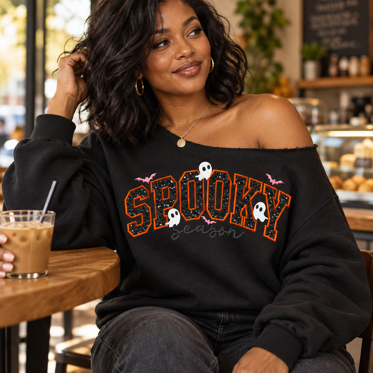 A woman sitting at a cafe table wearing a black off-the-shoulder sweatshirt with 'SPOOKY season' written in orange and white glitter-style lettering, with small ghost and bat graphics.
