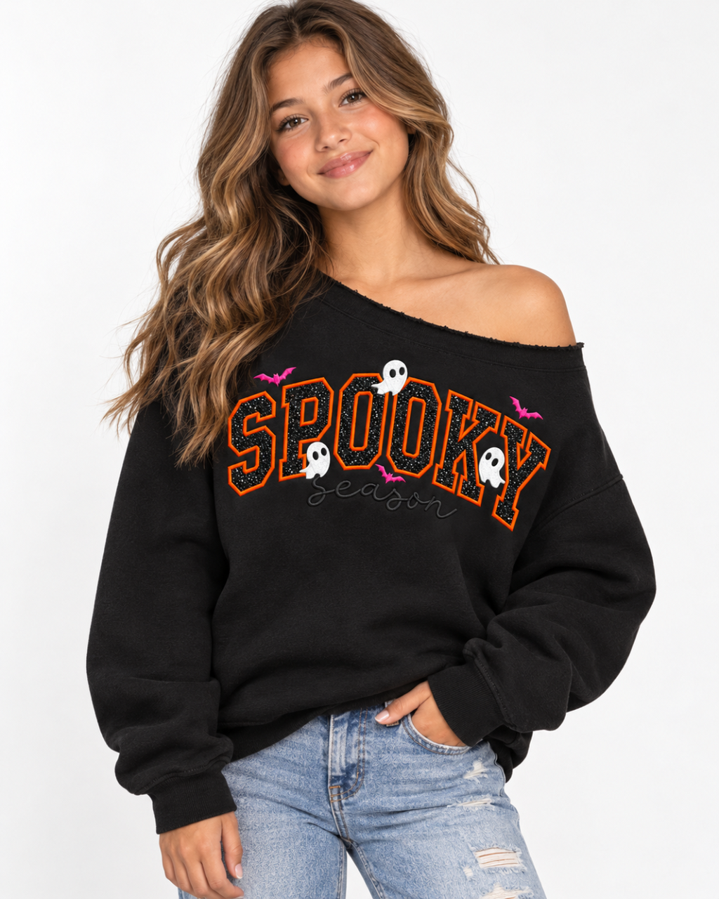 A woman wearing a black off-the-shoulder sweatshirt with 'SPOOKY season' written in orange outlined letters, decorated with white ghosts and pink bats, paired with light blue jeans against a white studio background.