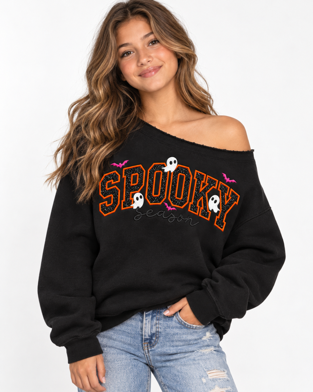 A woman wearing a black off-the-shoulder sweatshirt with 'SPOOKY season' written in orange outlined letters, decorated with white ghosts and pink bats, paired with light blue jeans against a white studio background.
