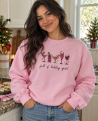 A woman wearing a light pink crewneck sweatshirt with a holiday-themed cocktail graphic and the text 'full of holiday spirit' in a kitchen setting.