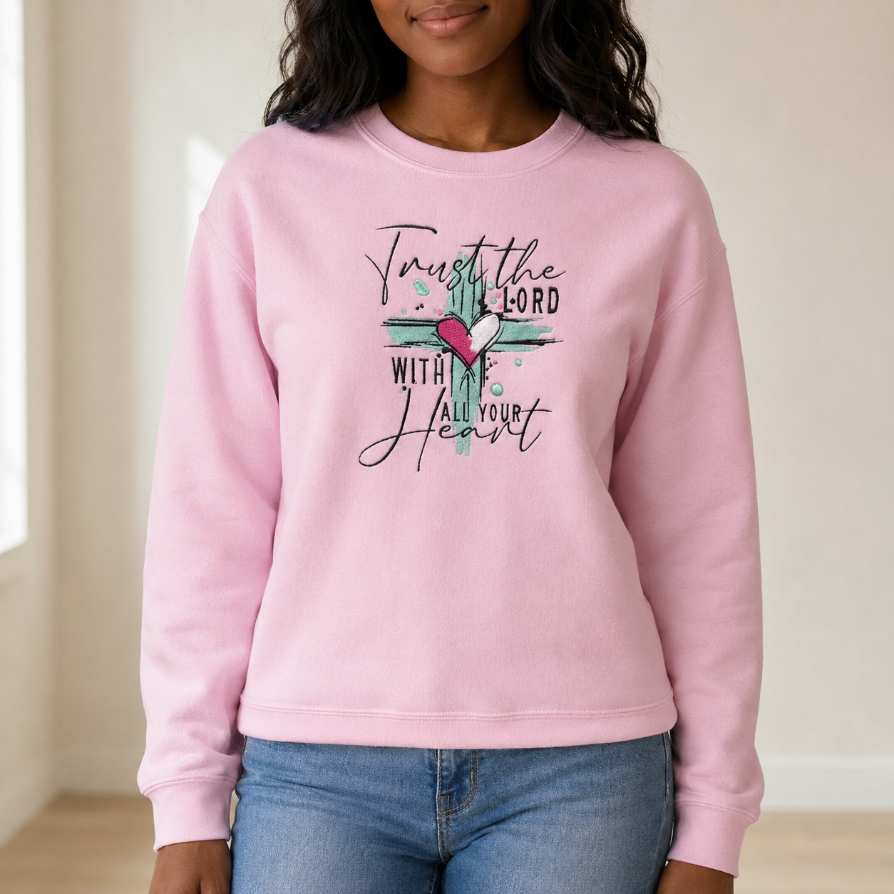 A woman wearing a light pink crewneck sweatshirt with a embroidered design the front that reads 'Trust the Lord with All Your Heart' in black script, featuring a teal cross and a pink heart. She is wearing blue jeans in a brightly lit indoor setting.