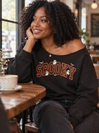 A woman with curly black hair sits at a wooden table in a cozy cafe, wearing a black off-the-shoulder sweatshirt with 'SPOOKY' written in orange letters and small white ghosts. A cup of coffee sits on the table next to her.