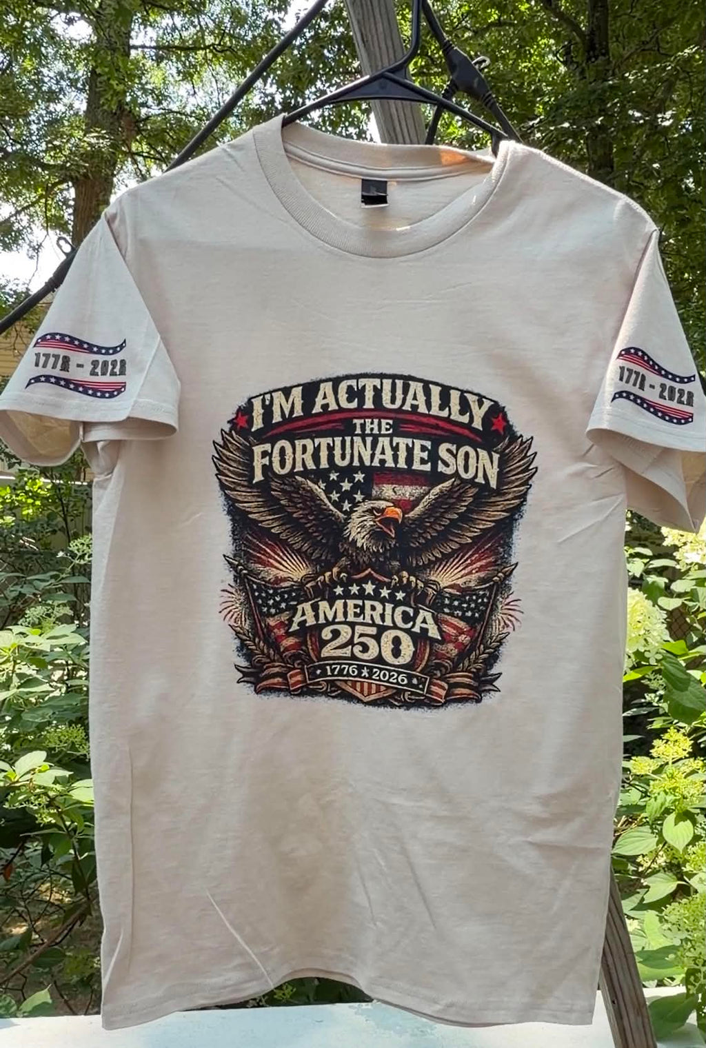 Actual shirt printed on a cream-colored short-sleeve t-shirt hanging on a black hanger outdoors against a background of green trees. The shirt features a large graphic print on the front with an eagle, stars, and the text 'I'M ACTUALLY THE FORTUNATE SON AMERICA 250'.
