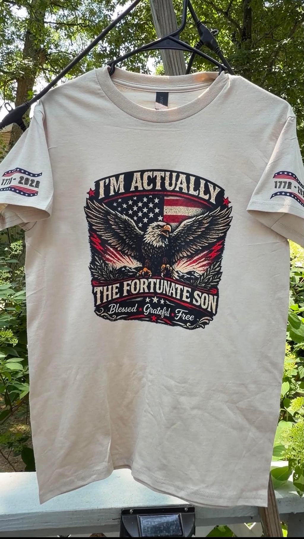 Actual shirt printed on cream-colored short-sleeve graphic t-shirt outdoors. The shirt features a large front graphic with an eagle, American flag, and the text 'I'M ACTUALLY THE FORTUNATE SON'.
