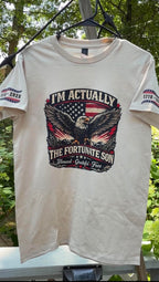 Actual shirt printed on cream-colored short-sleeve graphic t-shirt outdoors. The shirt features a large front graphic with an eagle, American flag, and the text 'I'M ACTUALLY THE FORTUNATE SON'.