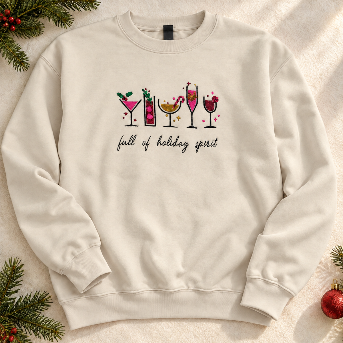 An tan crewneck sweatshirt with a festive graphic on the chest. The graphic using cocktail glasses filled with pink and yellow drinks, with the phrase 'full of holiday spirit' written below in black script.