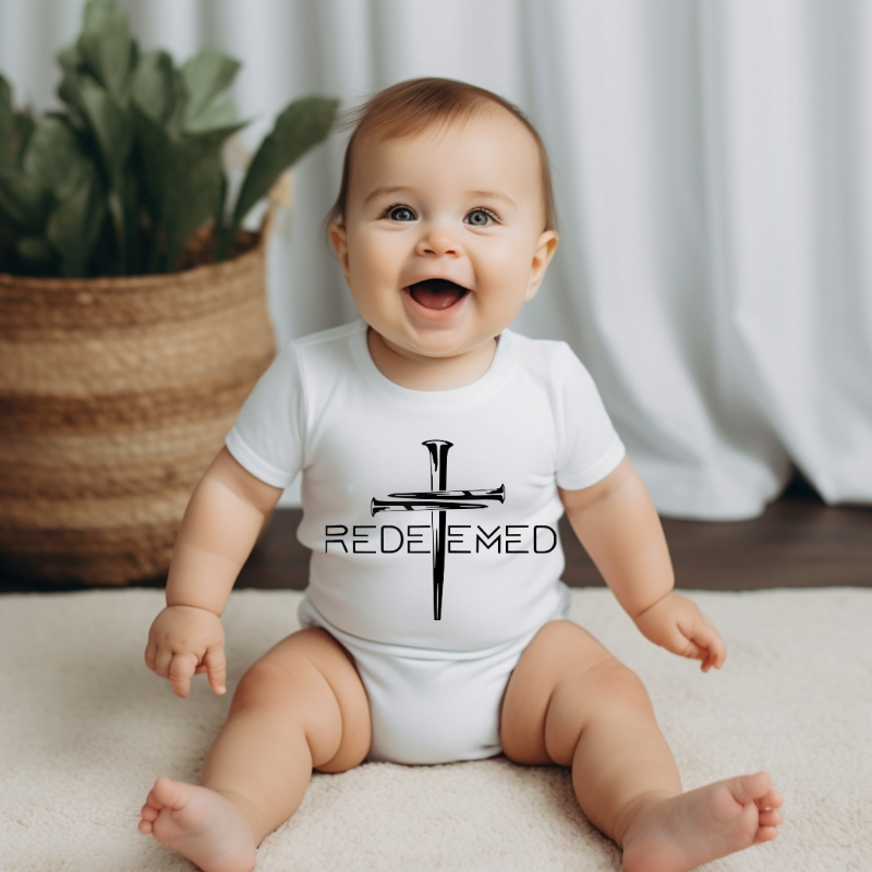 Baby wearing a white onesie with 'REDEEMED' text and cross design, sitting on a light-colored surface.