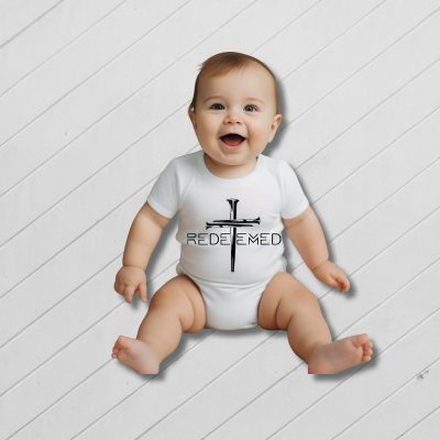  Baby wearing a white onesie with 'REDEEMED' text on a light wooden floor.