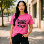 Woman wearing a pink t-shirt with 'Beautifully Broken' design outdoors.