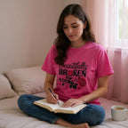 Woman in pink shirt with 'Beautifully Broken' design writing in a notebook on a bed.