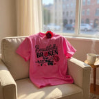 Pink t-shirt with 'Beautifully Broken' text and roses on a beige armchair near a window.