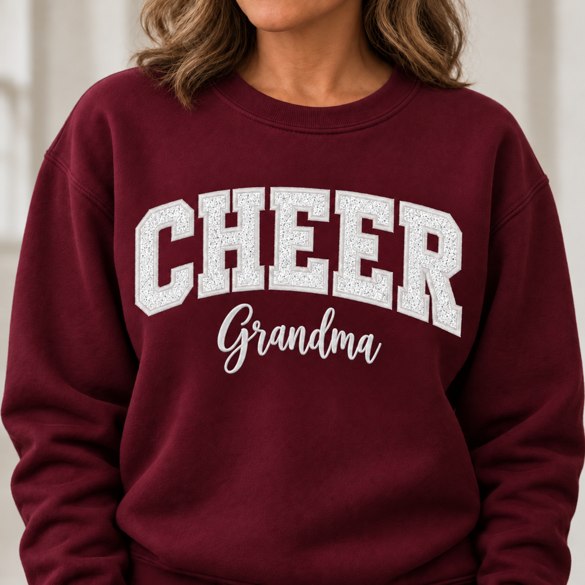 A close-up of a person wearing a burgundy crewneck sweatshirt with 'CHEER Grandma' printed in white across the chest.