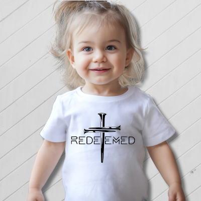 Child wearing a white t-shirt with a cross and 'REDEEMED' text on a light wooden floor.