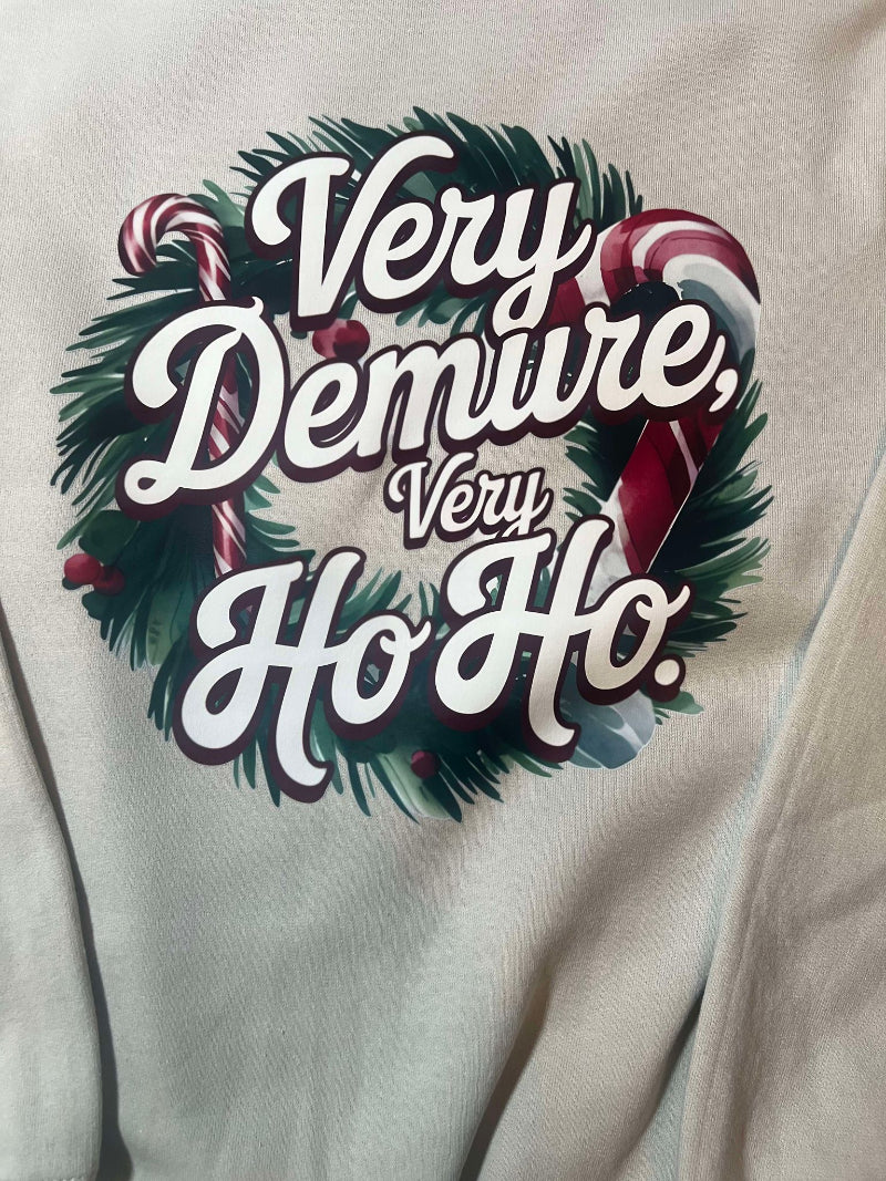 A cream-colored sweatshirt with 'Very Demure, Very Ho-Ho' text and a Christmas wreath design, displayed on a hanger.