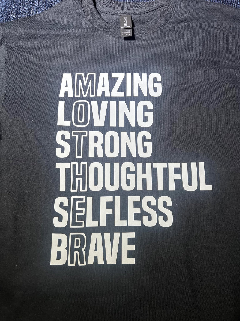 A black short-sleeve graphic tee with white text that lists adjectives like Amazing, Loving, Strong, and Brave, with the word 'MOTHER' highlighted vertically.