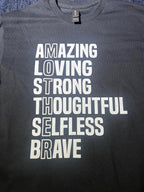 A black short-sleeve graphic tee with white text that lists adjectives like Amazing, Loving, Strong, and Brave, with the word 'MOTHER' highlighted vertically.