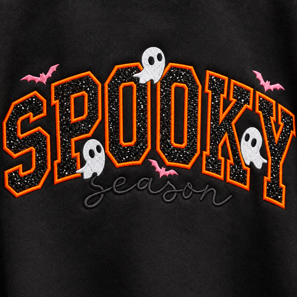 Close-up of a black garment featuring a large arched 'SPOOKY' graphic in black glitter-style fill with orange outlines, plus small white ghosts, pink bats, and the word 'season' in black script below.