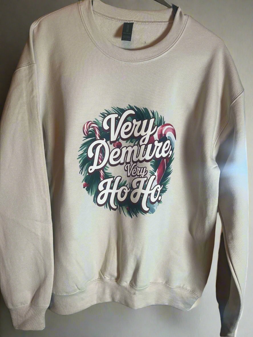 A cream-colored sweatshirt with 'Very Demure, Very Ho-Ho' text and a Christmas wreath design, displayed on a hanger.
