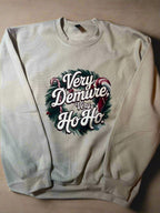 A cream-colored sweatshirt with 'Very Demure, Very Ho-Ho' text and a Christmas wreath design, displayed on a hanger.