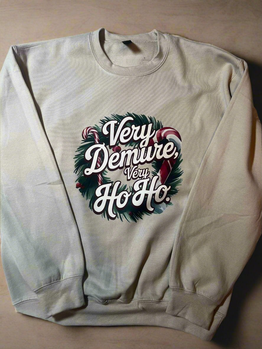 A cream-colored sweatshirt with 'Very Demure, Very Ho-Ho' text and a Christmas wreath design, displayed on a hanger.