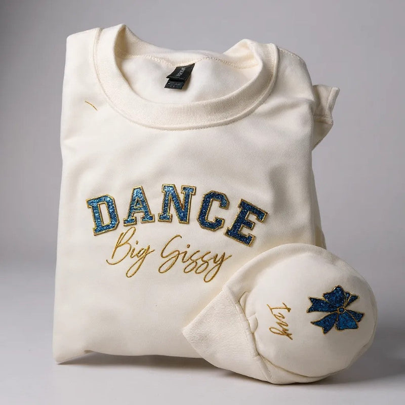 Folded white sweatshirt with 'DANCE Big Sissy' text and blue glitter applique  and gold stitch design on a gray background