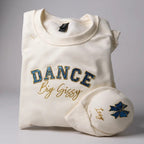 Folded white sweatshirt with 'DANCE Big Sissy' text and blue glitter applique  and gold stitch design on a gray background