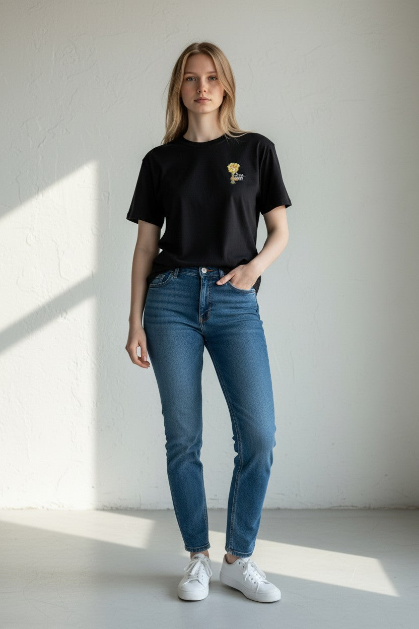 Woman wearing a black t-shirt with a logo and blue jeans standing against a white wall.