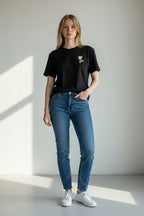 Woman wearing a black t-shirt with a logo and blue jeans standing against a white wall.