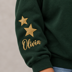 Close-up of a dark green sweatshirt sleeve with two gold glitter stars and the name 'Olivia' embroidered in gold script on the upper arm.