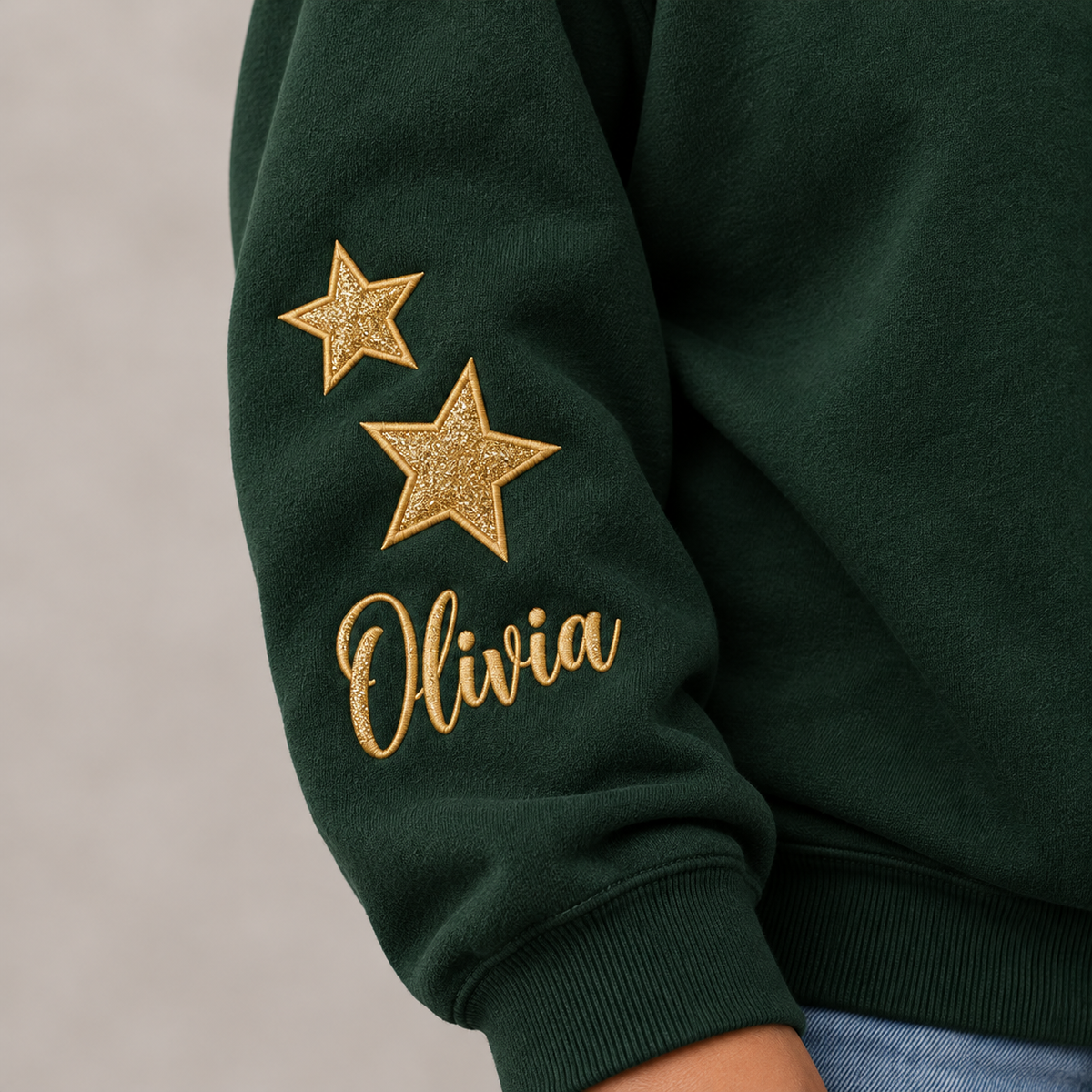 Close-up of a dark green sweatshirt sleeve with two gold glitter stars and the name 'Olivia' embroidered in gold script on the upper arm.