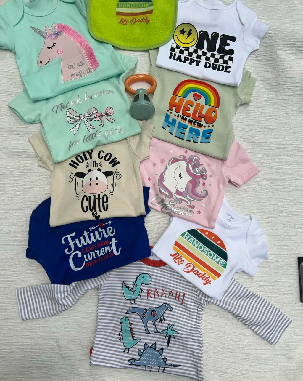 A collection of various baby tee shirts with different colors and graphics, including unicorn, smiley face, and dinosaur designs, along with text phrases such as 'future cowboy' and 'hippity hop'.