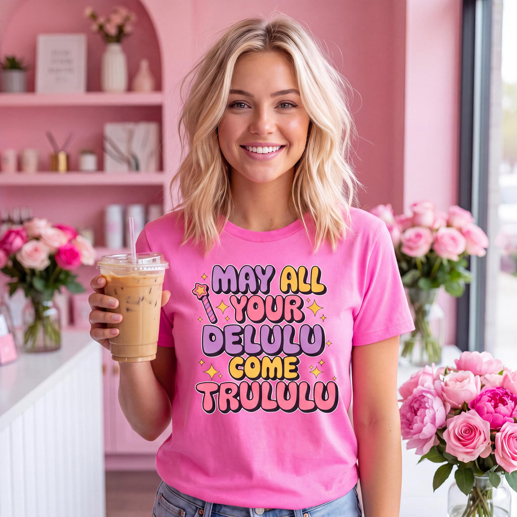A smiling blonde woman in a pink cafe setting holds an iced coffee and wears a bright pink graphic T-shirt with the text 'MAY ALL YOUR DELULU COME TRULULU' in colorful lettering.