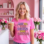 A smiling blonde woman in a pink cafe setting holds an iced coffee and wears a bright pink graphic T-shirt with the text 'MAY ALL YOUR DELULU COME TRULULU' in colorful lettering.