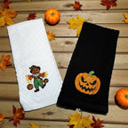 Two Halloween-themed towels, one white with a scarecrow and one black with a jack-o'-lantern, on a wooden surface with pumpkins and leaves.
