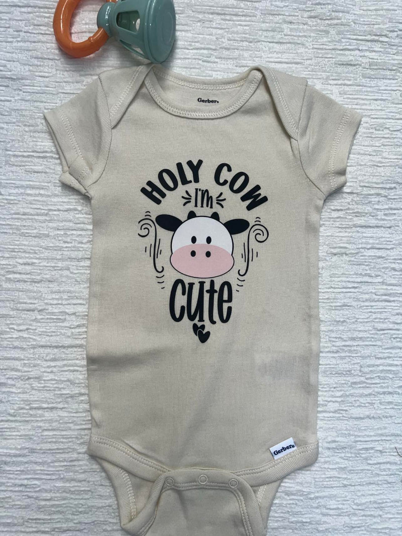 Beige baby onesie with a cow face and the words: "Holy Cow, I'm Cute"