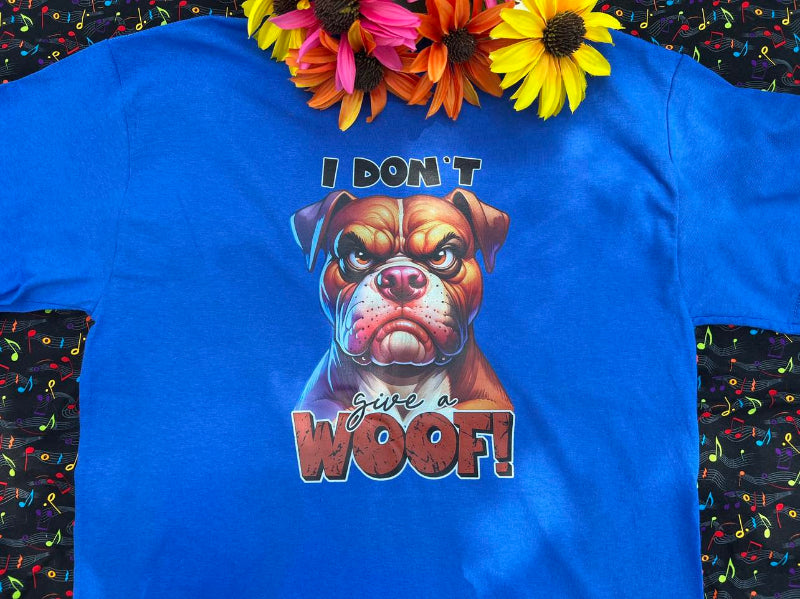 A Blue graphic t-shirt with a dog face and the words "I Don't Give A Woof" on a colorful background with flowers near the collar of shirt.