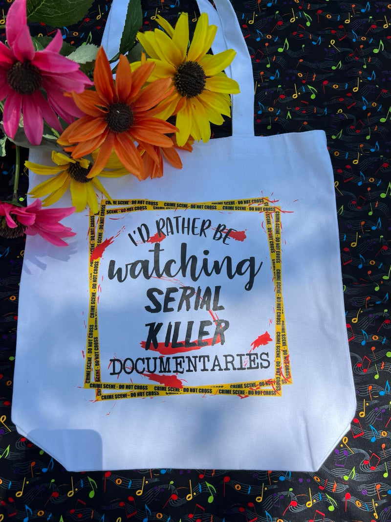 White canvas tote bag with the text 'I'd Rather Be Watching Serial Killer Documentaries' printed in black and yellow, accompanied by a decorative yellow crime scene tape.