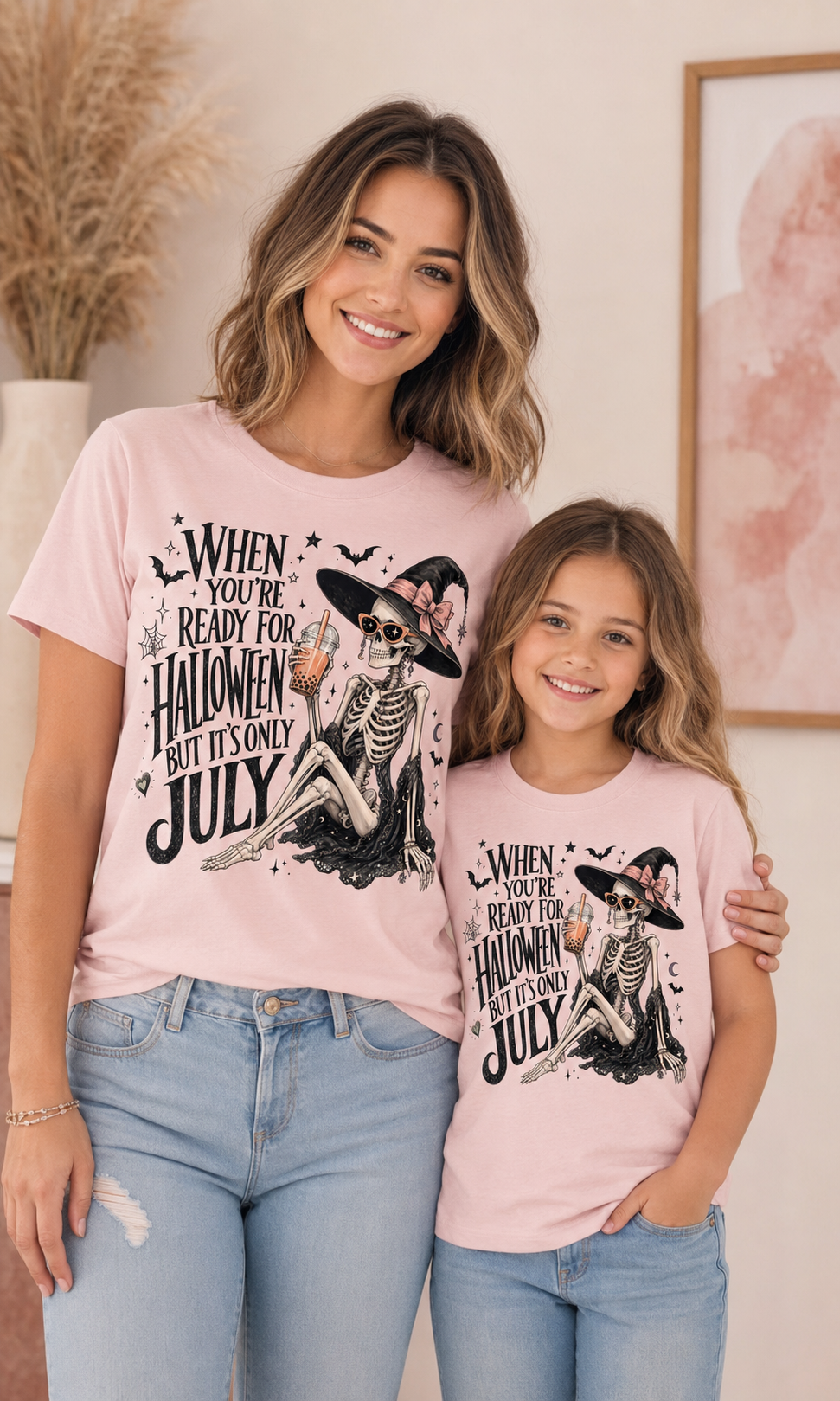 A woman and a child wearing matching light pink graphic t-shirts with a Halloween skeleton design and the text 'WHEN YOU'RE READY FOR HALLOWEEN BUT IT'S ONLY JULY'.
