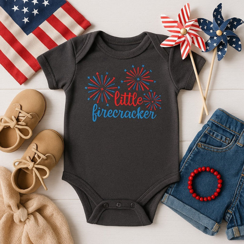 Black baby onesie with 'little firecracker' design, surrounded by American flag, shoes, denim shorts, and decorative items on a white surface.